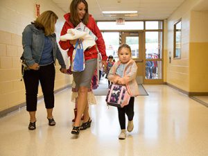 Student holding backpack walks down school hallway accompanied by teacher.