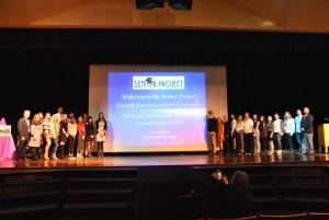 Two groups of participants in the Senior Project program pose on stage with advisors, teachers and administrators. A slide projection drop stands between two groups.