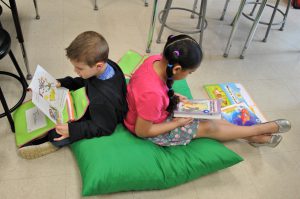 Two students sit back to bac on a large people reading picture books