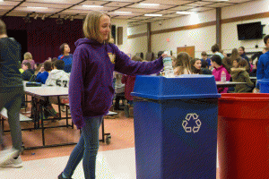 A girl in a purple sweatshirt drops a plastic water bottle into a recycle bin in the Middle School cafeteria
