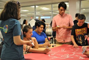 In the school's media center, a group of students sit or stand around a table. They are drawing a creature on red paper using tubes of toothpaste.on red paper.