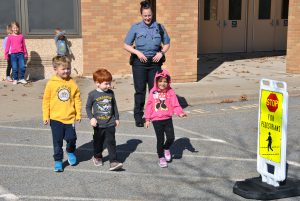 A school resource officer looks on as three kindergartners cross the street outside the school building.