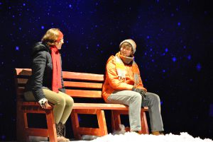 Two students dressed for the cold sit on a bench. There's a starry sky behind them and snow on the foreground.